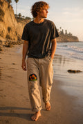 THC Shrooms at Night Patch displayed on the right thigh of well-worn tan canvas pants worn by a barefoot California beachgoer, featuring colorful embroidered mushrooms on a black half-circle background