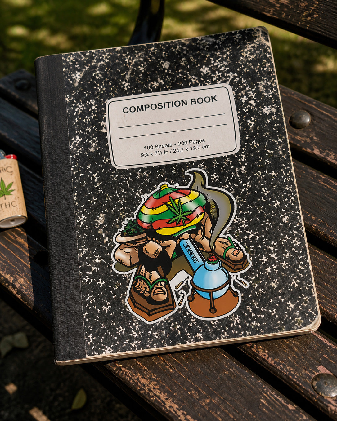 THC Rasta Joe sticker on black composition notebook on worn wooden park bench in outdoor setting