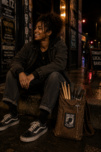 THC POW Patch displayed on the front flap of a worn brown canvas drumstick bag beside a young Black drummer sitting outside an independent Seattle music venue after a show featuring the End the Drug War embroidered graphic