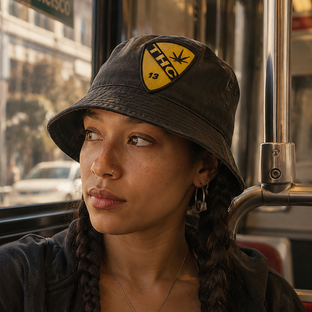 THC Air Cav Patch displayed on a faded black bucket hat worn by a young woman riding public transit in San Francisco featuring a yellow and black embroidered aviation-inspired shield with a cannabis leaf graphic