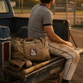 THC Basehit Patch displayed on the back pocket of dusty vintage baseball pants worn by a young man sitting on the tailgate of a weathered pickup beside a local baseball field featuring a red, white, and blue embroidered vintage baseball-inspired parody graphic