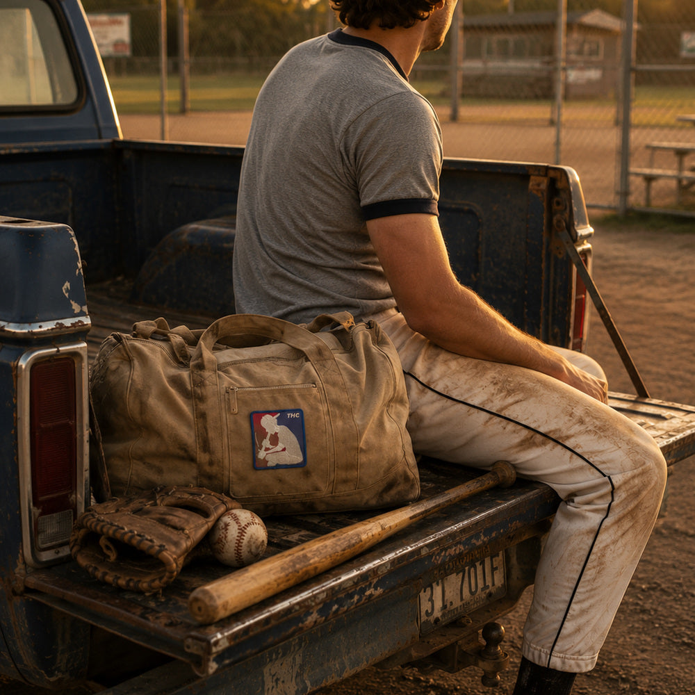 THC Basehit Patch displayed on the back pocket of dusty vintage baseball pants worn by a young man sitting on the tailgate of a weathered pickup beside a local baseball field featuring a red, white, and blue embroidered vintage baseball-inspired parody graphic