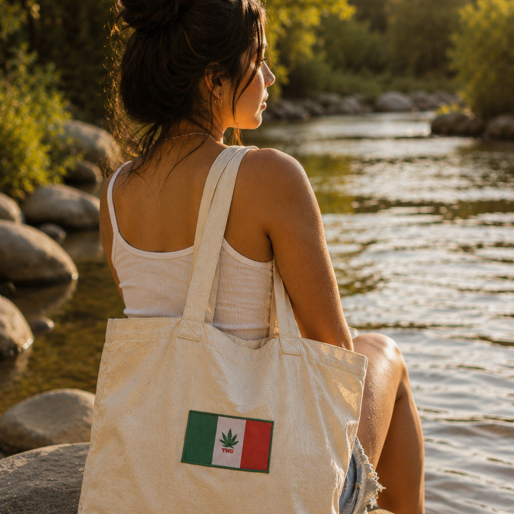 THC Mexico Flag Patch displayed on a natural canvas tote bag beside a young woman sitting on a riverside stone looking over a calm river featuring a green, white, and red embroidered Mexico-inspired flag with a cannabis leaf centerpiece