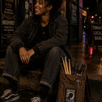 THC POW Patch displayed on the front flap of a worn brown canvas drumstick bag beside a young Black drummer sitting outside an independent Seattle music venue after a show featuring the End the Drug War embroidered graphic