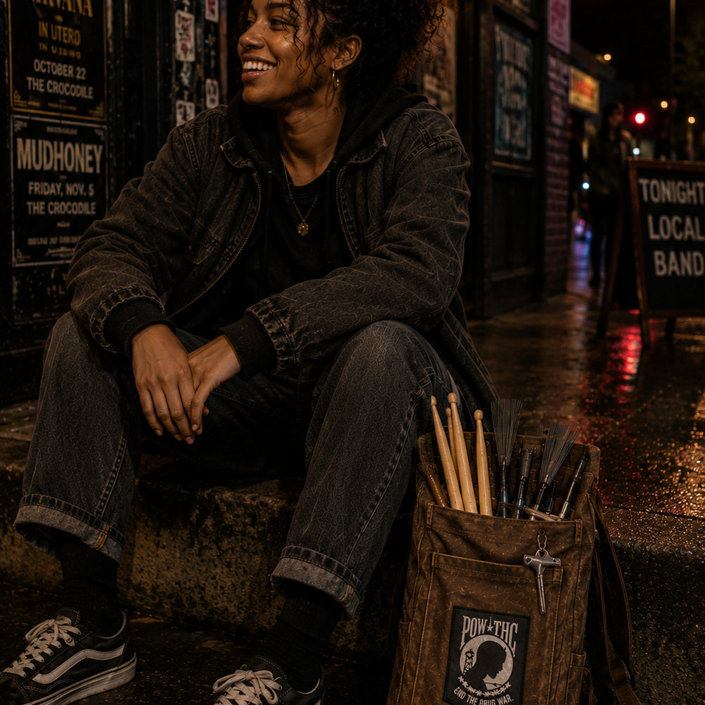 THC POW Patch displayed on the front flap of a worn brown canvas drumstick bag beside a young Black drummer sitting outside an independent Seattle music venue after a show featuring the End the Drug War embroidered graphic