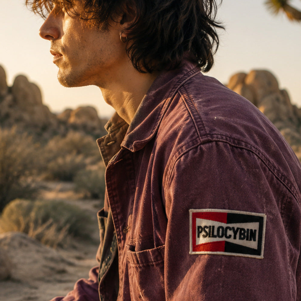 THC Psilocybin Patch displayed on the upper sleeve of a faded plum canvas chore jacket worn by a young artist in Joshua Tree at golden hour, featuring a retro embroidered psilocybin graphic in red, black, and white