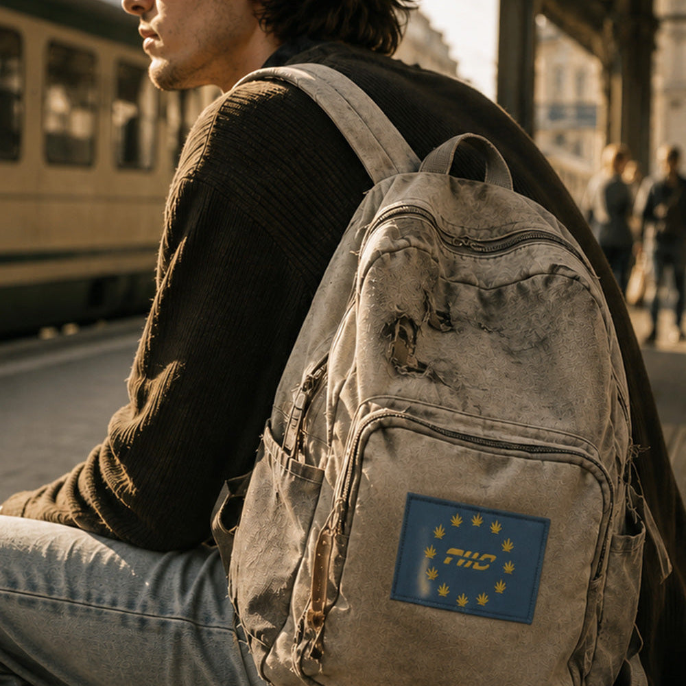 THC Europaz Patch displayed on the front pocket of a weathered light gray canvas backpack carried by a young traveler at a European train station, featuring a blue embroidered flag design with gold cannabis leaves stitched directly into the fabric