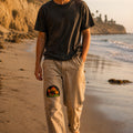 THC Shrooms at Night Patch displayed on the right thigh of well-worn tan canvas pants worn by a barefoot California beachgoer, featuring colorful embroidered mushrooms on a black half-circle background