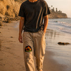 THC Shrooms at Night Patch displayed on the right thigh of well-worn tan canvas pants worn by a barefoot California beachgoer, featuring colorful embroidered mushrooms on a black half-circle background