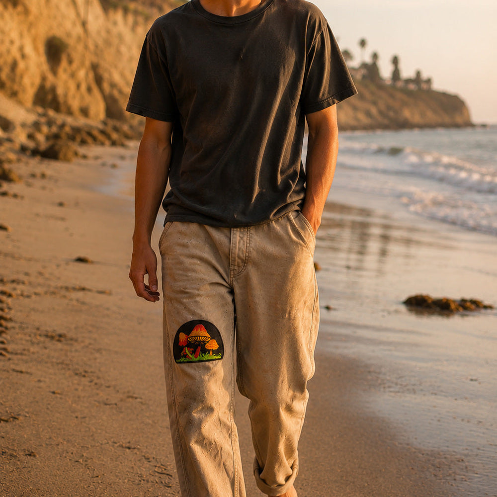 THC Shrooms at Night Patch displayed on the right thigh of well-worn tan canvas pants worn by a barefoot California beachgoer, featuring colorful embroidered mushrooms on a black half-circle background