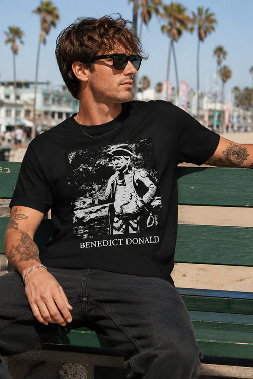 Man wearing a black t-shirt with a graphic design sitting on a bench with palm trees in the background.